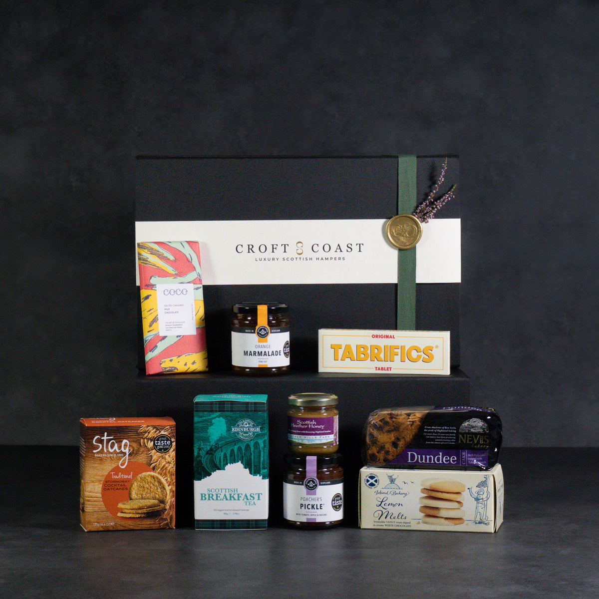 A curated gourmet gift hamper from Croft & Coast, featuring a black presentation box with a white label and green ribbon, surrounded by various Scottish snacks and treats including Stagg biscuits, Marcellus jam, Pickle, Dundee cheese, and a breakfast box, set against a dark textured background.