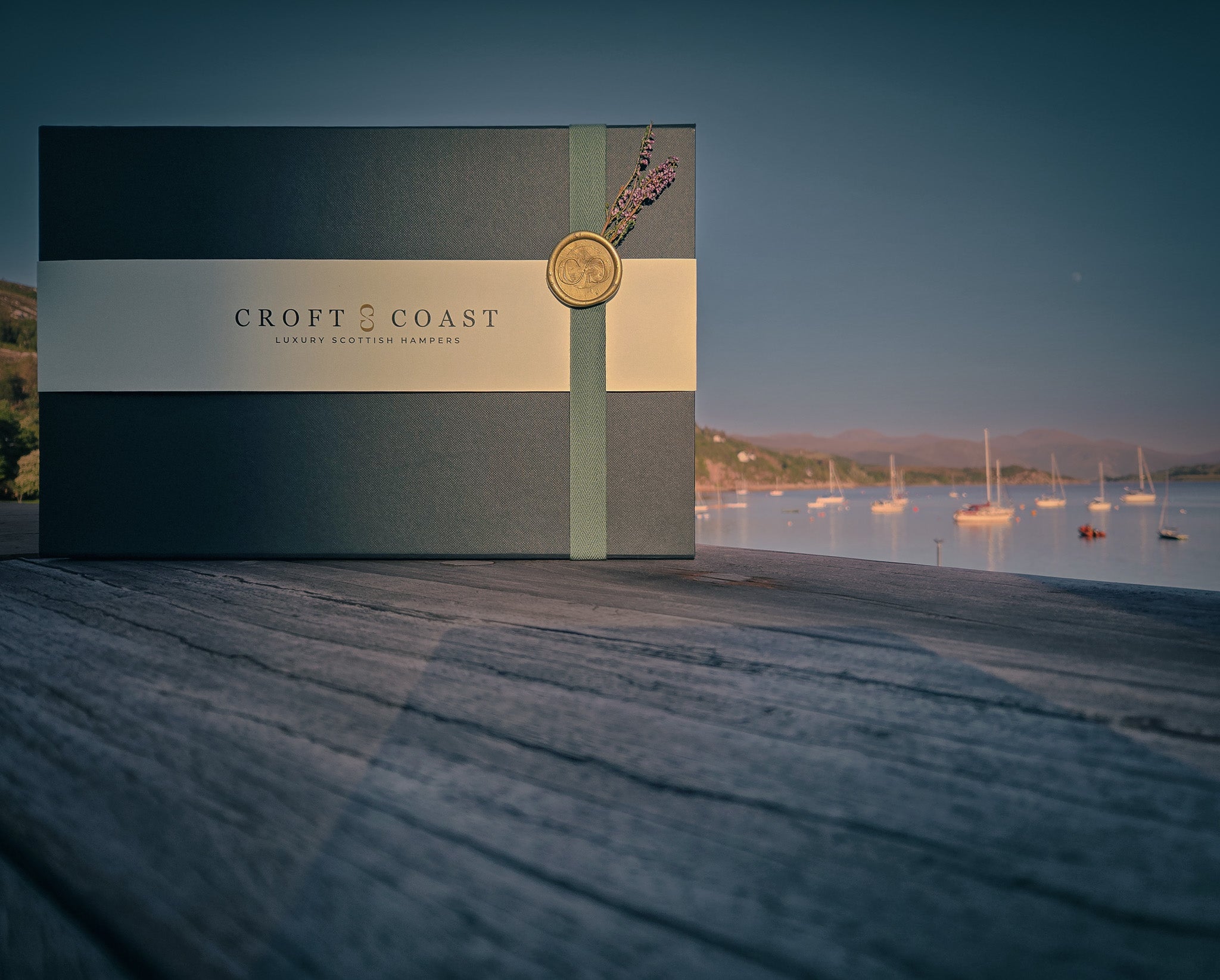 A Croft & Coast luxury gift hamper featuring a signature black presentation box, belly band, and green ribbon with wax seal, resting on a wooden surface with a scenic, sunlit view of boats at Ullapool Harbour in the background.