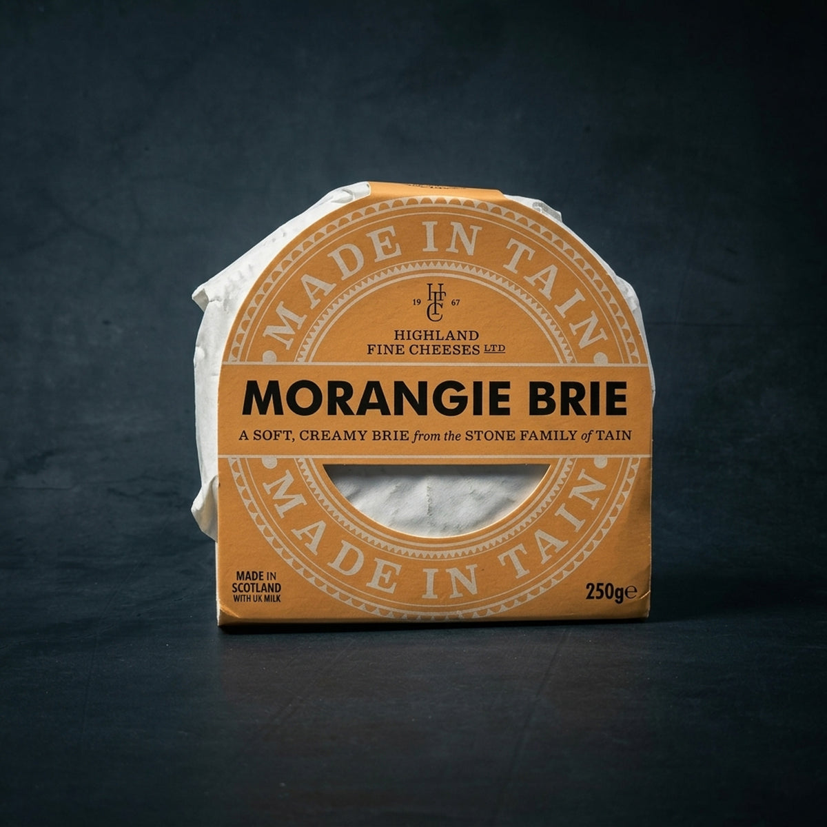 A wheel of Morangie Brie by Highland Fine Cheeses, featuring its signature tan and cream "Made in Tain" packaging, displayed on a dark grey textured surface.