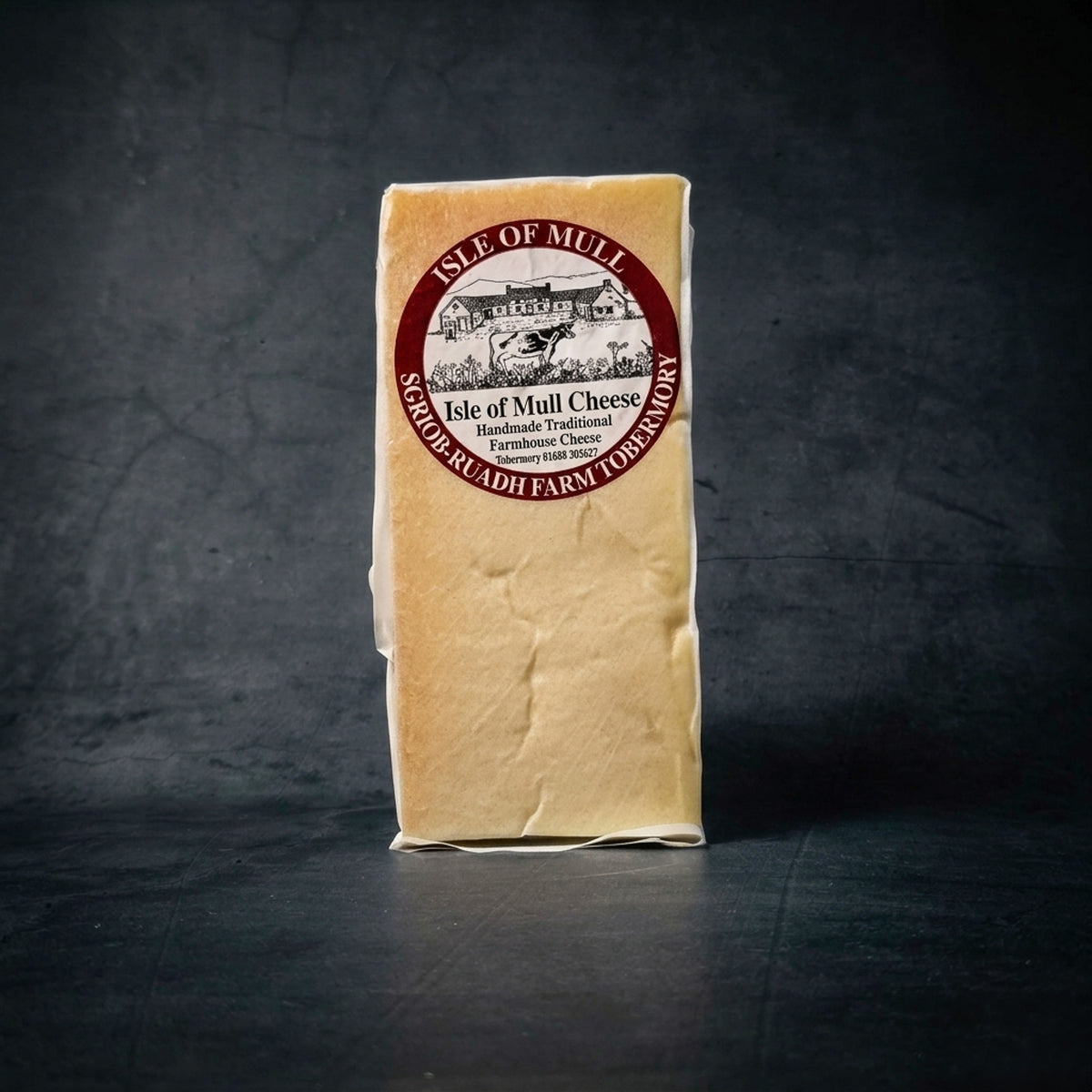 A block of Isle of Mull Traditional Farmhouse Cheese from Sgriob-ruadh Farm, Tobermory, in its clear protective wrapping with a distinctive circular label, displayed on a dark grey textured surface.