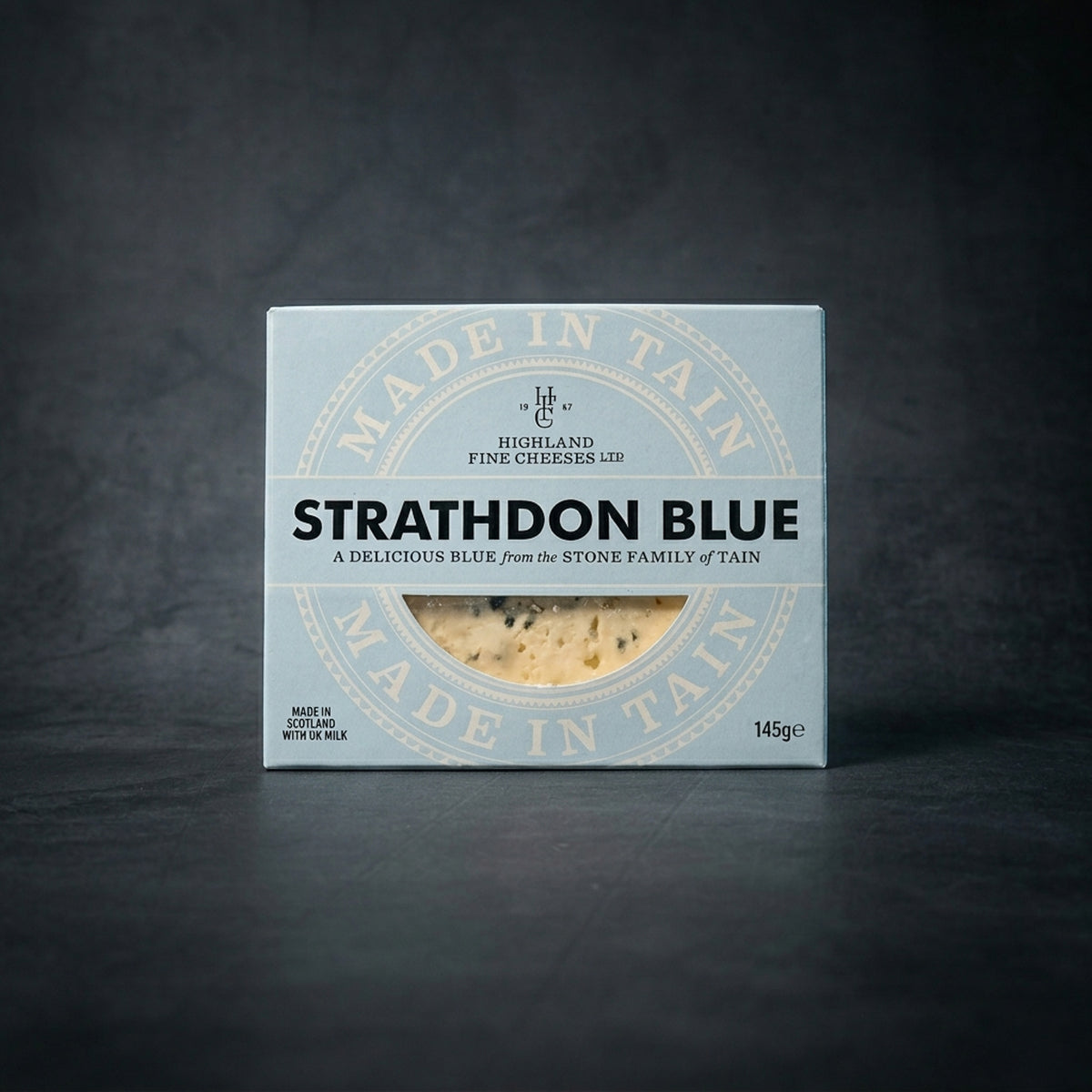 A box of Strathdon Blue cheese by Highland Fine Cheeses, featuring its signature blue and white branding, displayed on a dark grey textured surface.