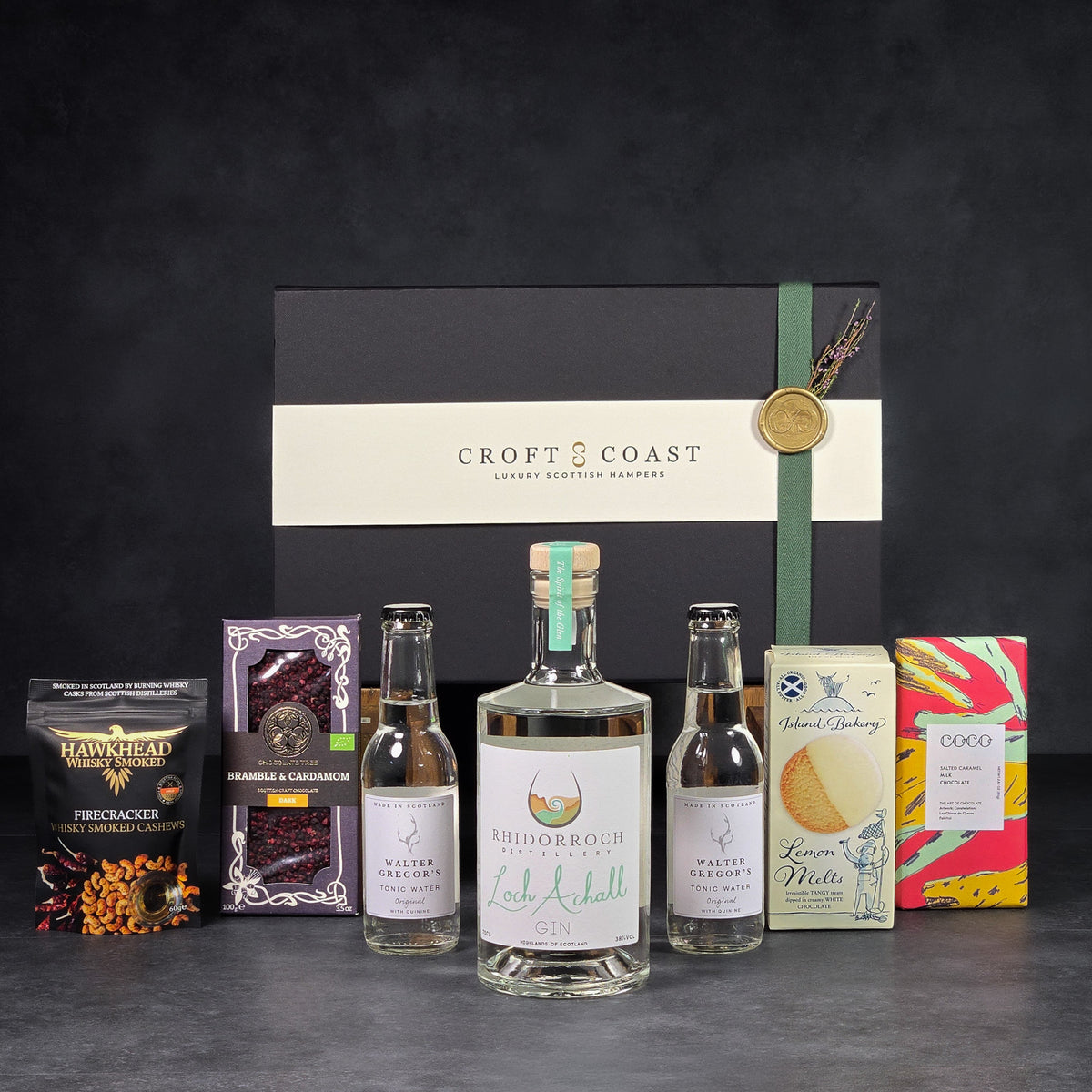 The Welcome Toast hamper featuring a signature black presentation box displayed behind a bag of smoked cashews, two chocolate bars, a bottle of gin, two small bottles of tonic water, and a box of lemon melt biscuits arranged on a dark grey textured surface.